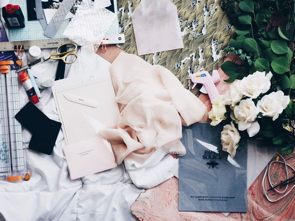 Mesa de trabajo con telas en tonos crema y rosa, un cuaderno, tijeras, regla y flores blancas