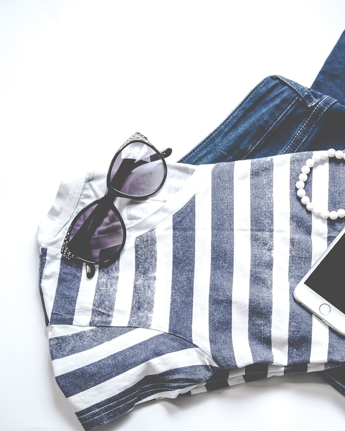 Camiseta de rayas, vaqueros, gafas de sol y pulsera de perlas ordenados sobre un fondo blanco